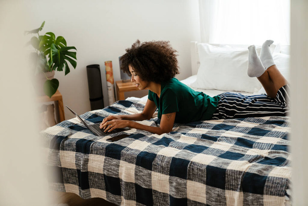 African american young woman with curly hairstyle lying in bed and using laptop at home - Foto, Imagem