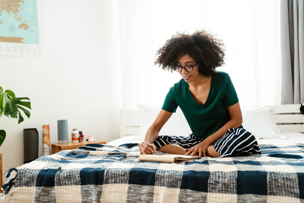 African american young woman with curly hairstyle sitting in bed and drawing in sketchbook at home - Photo, image