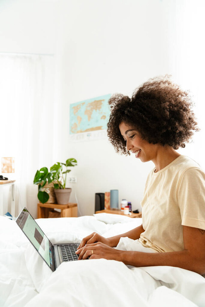 African american young woman with curly hairstyle lying in bed and using laptop at home - Photo, Image