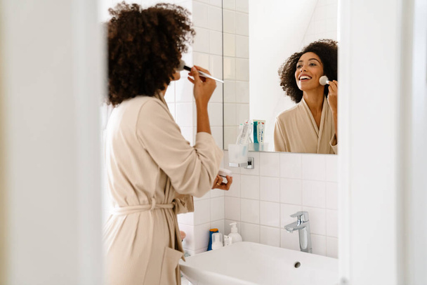 Junge Afroamerikanerin blickt in Spiegel und schminkt sich zu Hause mit Kosmetikpinsel - Foto, Bild