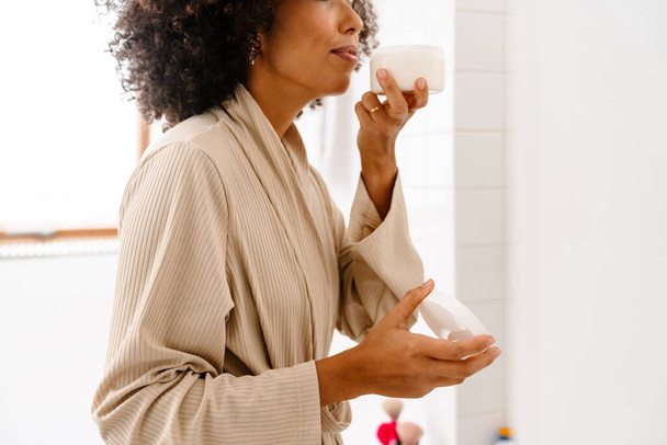 African american young woman with curly hairstyle smiling and holding face cream in bathroom - Foto, afbeelding