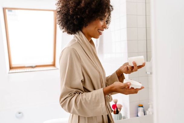 African american young woman with curly hairstyle smiling and holding face cream in bathroom - Photo, Image