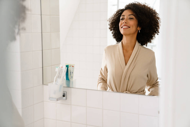 African american young woman with curly hairstyle smiling and looking in mirror at bathroom - 写真・画像