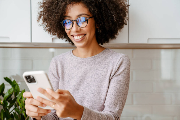 African american young woman with curly afro hairstyle smiling while using cellphone at home - Photo, Image