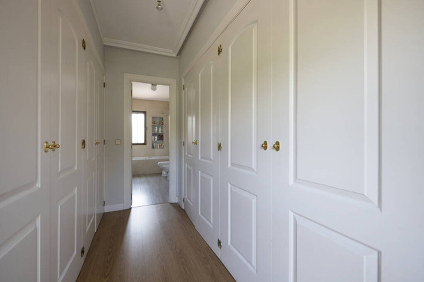 Habitación vacía con armarios empotrados de madera lacada en blanco formando un pasillo vestidor con acceso a un baño en suite - Foto, Imagen