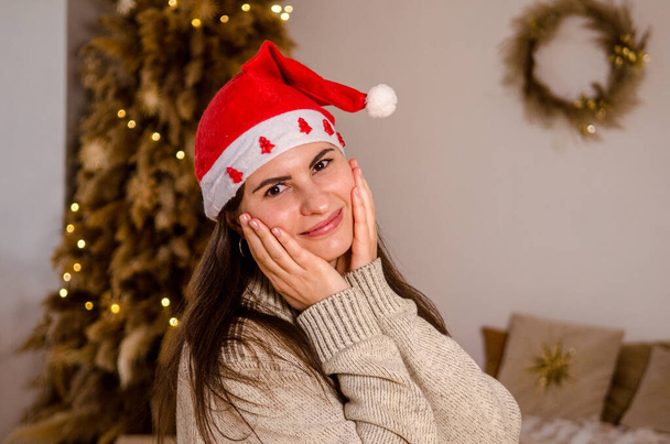 Mulher bonita nova em um suéter e com cabelo escuro em um chapéu vermelho de Papai Noel com estrelas, no fundo de uma cama e uma árvore de Natal em um quarto brilhante, feliz segurando seu rosto com as mãos - Foto, Imagem