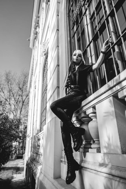 Modèle jeune et skinny avec de longs cheveux blonds et noirs et des vêtements et des bottes noires posant à l'extérieur de la fenêtre d'un bâtiment vieux siècles (en noir et blanc) - Photo, image