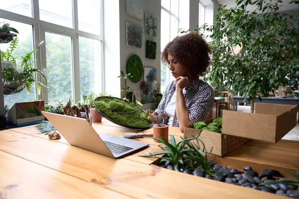 Young female is sitting at desk with laptop and holding plant for delivering it customers - Photo, Image