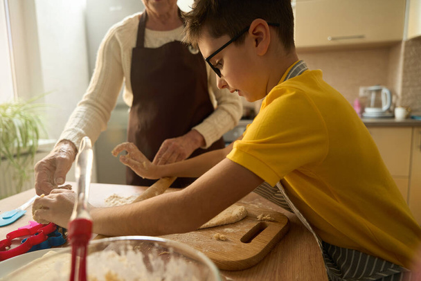 Boy standing near table in kitchen, rolls out dough with rolling pin, grandmother helps nearby - Photo, Image
