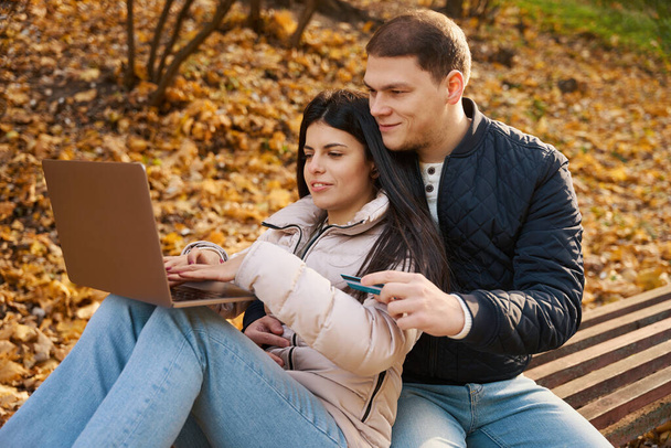 Young couple is browsing something in a laptop, they are located on a park bench - Photo, image
