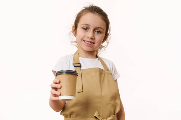 Gyönyörű kék szemű fehér kislány bézs szakácskötényt visel, forró italt, teát vagy kávét osztogat egy környezetbarát papírpohárban, aranyos mosollyal a kamerába néz. - Fotó, kép