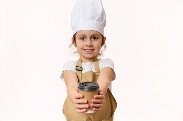 Καυκάσιο κοριτσάκι ντυμένο μπαρίστα, μπάρμαν με λευκό καπέλο σεφ και ποδιά, χαμογελώντας με ένα όμορφο χαμόγελο και δίνοντας στην κάμερα ένα ζεστό ρόφημα σε οικολογικό χάρτινο κύπελλο για να πάει, απομονωμένο σε λευκό - Φωτογραφία, εικόνα