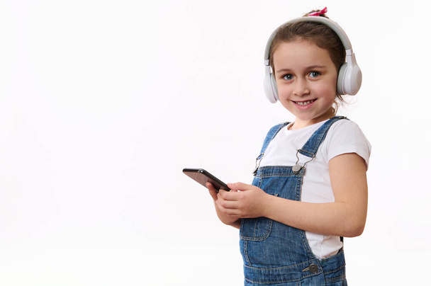Izolovaný portrét na bílém pozadí kavkazského chytrého dítěte, roztomilé holčičky v džínových montérkách, s bezdrátovými sluchátky, s mobilním telefonem a roztomile se usmívajícími pohledy do kamery - Fotografie, Obrázek