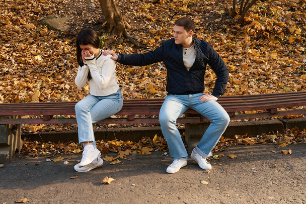 Young guy comforts his crying girlfriend, they are sitting on a park bench - Zdjęcie, obraz