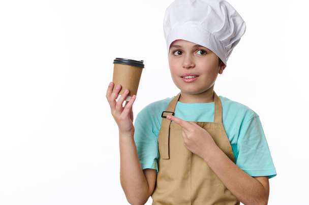 Hispanische hübsche Preteen Boy als Barkeeper Barista in beige Kochschürze und Hut gekleidet, zeigt auf einen zum Mitnehmen heißen Kaffee Teegetränk in Öko-Pappbecher, isoliert auf weißem Hintergrund. Freie Werbefläche - Foto, Bild