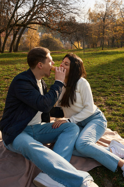 Cheerful young couple sits on the lawn in the city park, the guys treat themselves to sweets - Zdjęcie, obraz