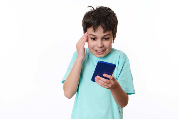 Surprised overjoyed multi-ethnic teenager, schoolboy wearing blue t-shirt, holding his hand on his temple, expressing astonishment while reading news on his smartphone, isolated over white background - Photo, Image