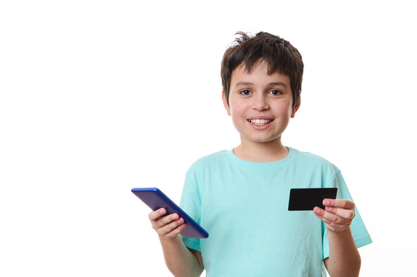 Привабливий чорноволосий кавказький хлопчик-підліток, розумний школяр у блакитній футболці зі смартфоном та пластиковою кредитною карткою, ізольований на білому тлі. Інтернет-банкінг. Безготівкова оплата. Інтернет - шопінги - Фото, зображення