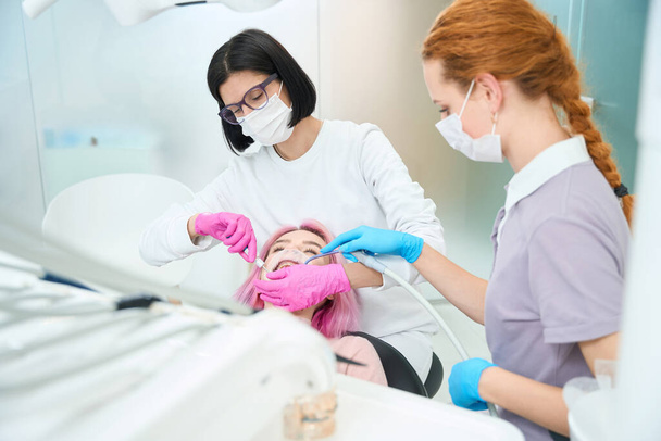 Жінка-стоматолог проводить стоматологічну процедуру молодому пацієнту, асистент допомагає лікарю
 - Фото, зображення