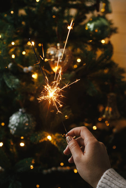 Hand holding firework against christmas tree lights in dark room. Happy New Year! Merry Christmas! Burning sparkler in female hand  on background of golden illumination bokeh. Atmospheric time - Photo, Image