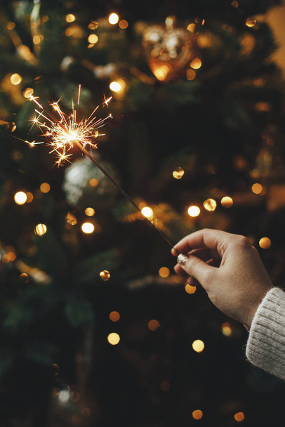 Hand holding firework against christmas tree lights in dark room. Happy New Year! Merry Christmas! Burning sparkler in female hand  on background of golden illumination bokeh. Atmospheric time - Photo, Image