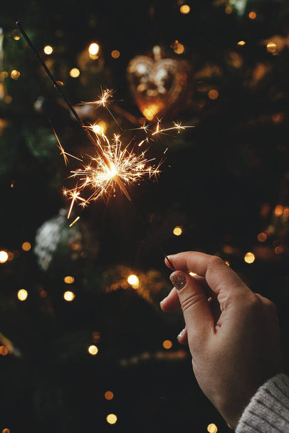 Brandende ster in vrouwelijke hand op de achtergrond van kerstboom lichten in donkere kamer. Gelukkig Nieuwjaar! Hand houden van vuurwerk tegen stijlvolle versierde boom met verlichting. Atmosferische tijd - Foto, afbeelding