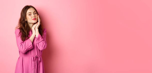 Beautiful and tender woman touching her perfect hydrated face with bright make up, holding hand on cheek, gazing at camera, standing over pink background. - Photo, Image