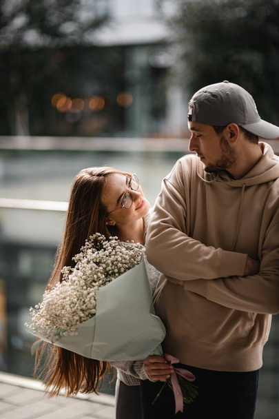 young smiling woman with long hair and bouquet of white flowers in her hands looks at man with cap. Couple in love. Love story - Foto, afbeelding