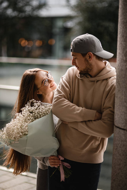 young smiling woman with bouquet of white flowers in her hands looks at man with cap. Couple in love. Love story - 写真・画像