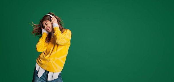 Portrait of young beautiful girl posing, emotionally listening to music in headphones isolated over green background. Concept of youth, beauty, fashion, lifestyle, emotions, facial expression. Ad - Zdjęcie, obraz