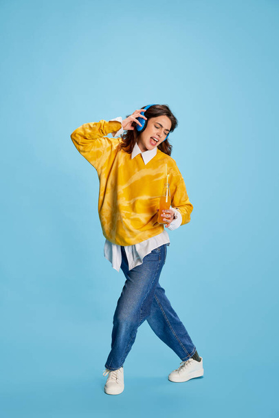 Portrait of young beautiful girl in casual clothes listening to music in headphones isolated over blue background. Concept of youth, beauty, fashion, lifestyle, emotions, facial expression. Ad - 写真・画像