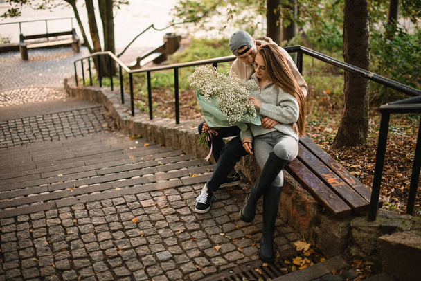 Attractive young woman and man sitting on bench outdoor. Couple in love hugs and kisses. Guy kisses girlfriend. Love story. - Fotografie, Obrázek