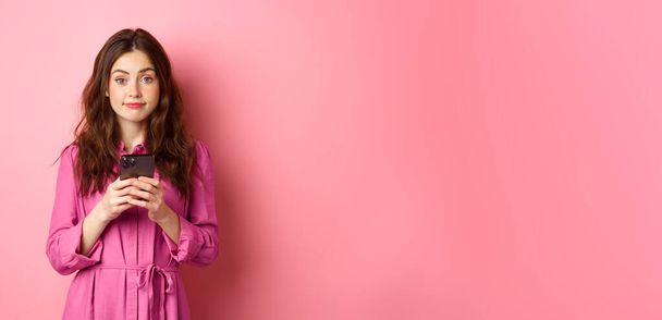 Cute young woman using mobile phone app, holding smartphone and smiling at camera, standing in dress against pink background. Copy space - Foto, Imagem