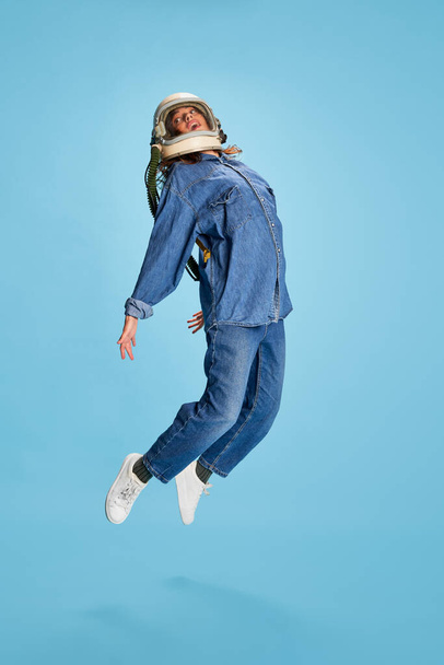 Portrait of young beautiful girl posing in uniform and astronaut helmet isolated over blue background. Jumping. Concept of youth, beauty, fashion, lifestyle, emotions, facial expression. Ad - Fotografie, Obrázek