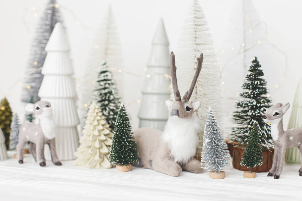 メリークリスマス！白い背景にスタイリッシュな小さなクリスマスツリーとトナカイのおもちゃ。お祭りのクリスマスシーン、鹿とミニチュアの雪の森。モダンな装飾、休日のバナー - 写真・画像