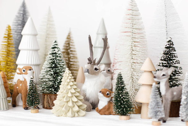 Merry Christmas! Stylish little Christmas trees and reindeer toys on white background. Festive Christmas scene, miniature snowy forest with cute deers. Modern decorations, holiday banner - Photo, Image