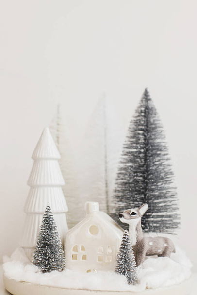 Vrolijk kerstfeest! Stijlvolle kerstboompjes en rendierspeelgoed op witte tafel. Kerstsprookjesscène, miniatuur besneeuwd bos met schattig hert. Moderne tafelopstelling, monochromatische decoraties - Foto, afbeelding
