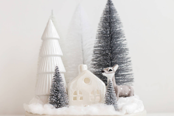 Pequenas árvores de Natal elegantes e brinquedo de rena na mesa branca. Cena festiva de Natal, floresta nevada em miniatura com cervos bonitos. Feliz Natal! Cenário de mesa moderno, decorações monocromáticas - Foto, Imagem