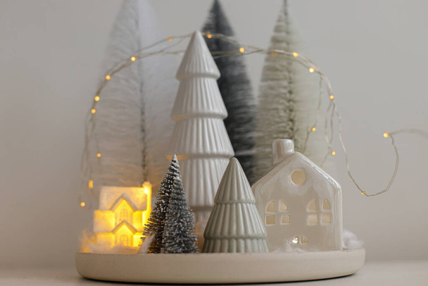 Χειμώνας hygge, ζεστό Χριστούγεννα μαγική σκηνή, μινιατούρα χιονισμένο χωριό με φώτα. Κομψά χριστουγεννιάτικα δέντρα και σπίτι σε λευκό τραπέζι. Καλά Χριστούγεννα! Μοντέρνα διακόσμηση διακοπών, ατμοσφαιρικός χρόνος - Φωτογραφία, εικόνα