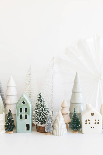 Hyvää joulua! Tyylikäs pikku joulukuusia ja taloja koristeet valkoisella pöydällä. Moderni joulu kohtaus, miniatyyri viihtyisä luminen kylä. Talviloma banneri, skandaali sisustus - Valokuva, kuva