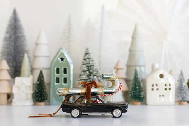 Holidays are coming! Stylish little car carrying candy cane on background of Christmas miniature snowy village. Merry Christmas! Festive winter scene on white table, xmas banner - Photo, Image