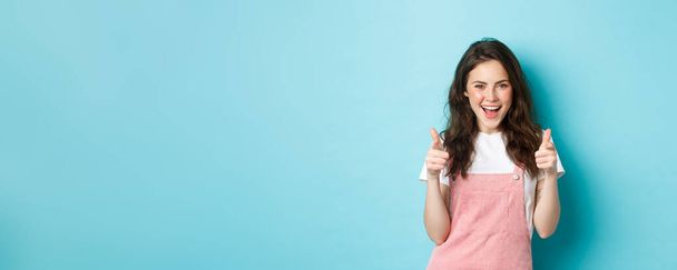 Confident and cheerful brunette girl say yes, pointing fingers at camera and looking sassy, praising you, inviting to event, standing over blue background. - Foto, Imagen