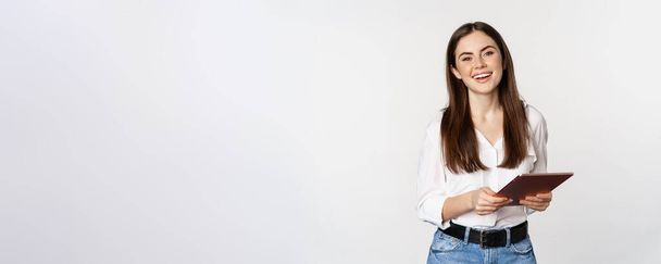 Смолящая современная женщина, стоящая с цифровой табличкой, смеющаяся и выглядящая счастливой, работающая, позирующая на белом студийном фоне. - Фото, изображение