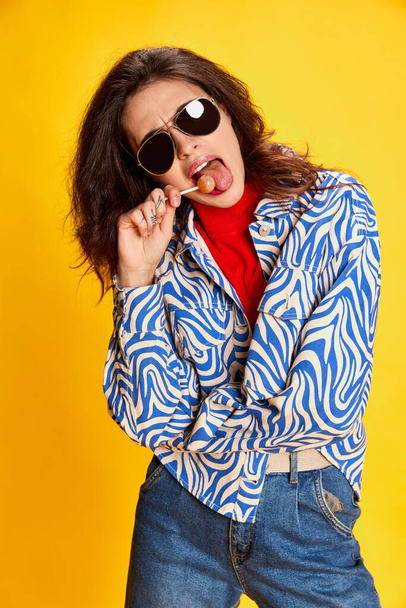 Portrait of young beautiful girl posing in stylish clothes and sunglasses, eating lollipop isolated on yellow background. Concept of youth, beauty, fashion, lifestyle, emotions, facial expression. Ad - Photo, Image