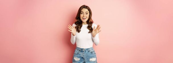 Image of excited smiling stylish woman, shaking hands and jumping enthusiastic, looking amazed at camera, standing against pink background. - Fotografie, Obrázek