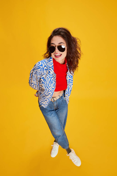 Portrait of young beautiful girl posing in vintage clothes, eating lollipop isolated over yellow background. Concept of youth, beauty, fashion, lifestyle, emotions, facial expression. Ad - Foto, afbeelding