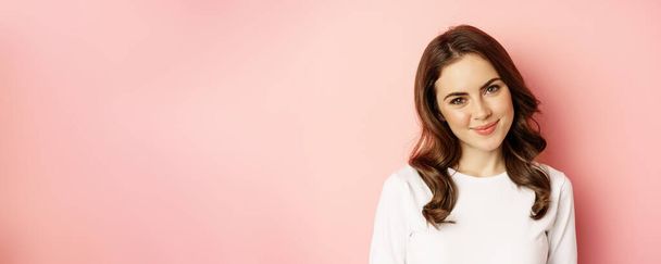 Close up of enthusiastic brunette girl with makeup, smiling and looking happy at camera, posing against pink background. - Фото, зображення