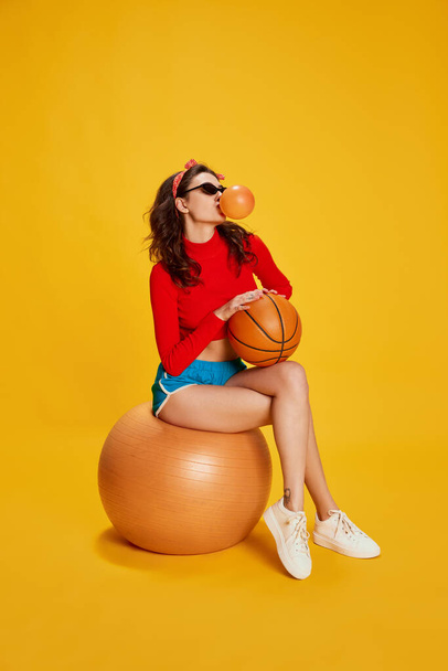Portrait of young stylish girl posing, sitting on rubber fitness ball isolated over yellow background. Bubble gum. Concept of youth, beauty, sport lifestyle, emotions, facial expression. Ad - Photo, image