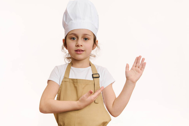 Delighted baby girl dressed as pastry chef or baker confectioner, in chefs hat and apron, pointing at copy space for your promotional text on white background. Concept of bakery, culinary, cookery - Fotografie, Obrázek