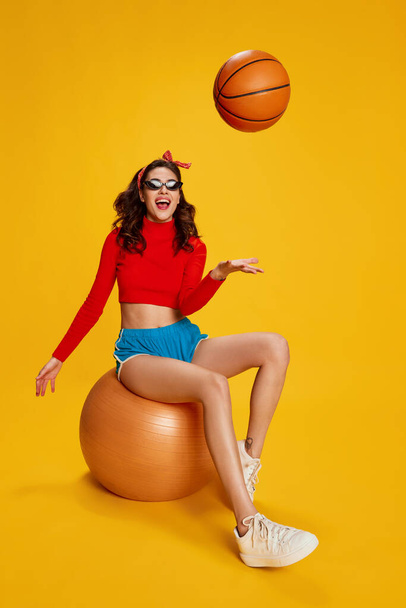 Portrait of young beautiful girl posing, sitting on rubber fitness ball isolated over yellow background. Gym games. Concept of youth, beauty, sport lifestyle, emotions, facial expression. Ad - 写真・画像